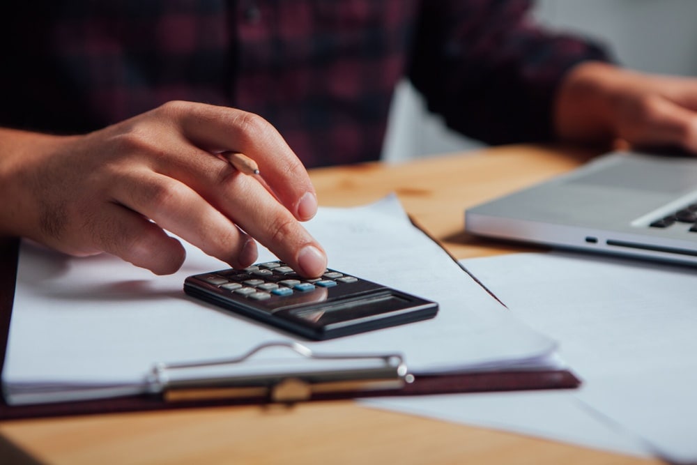O mână folosește un calculator peste niște documente, cu un laptop neclar în fundal.