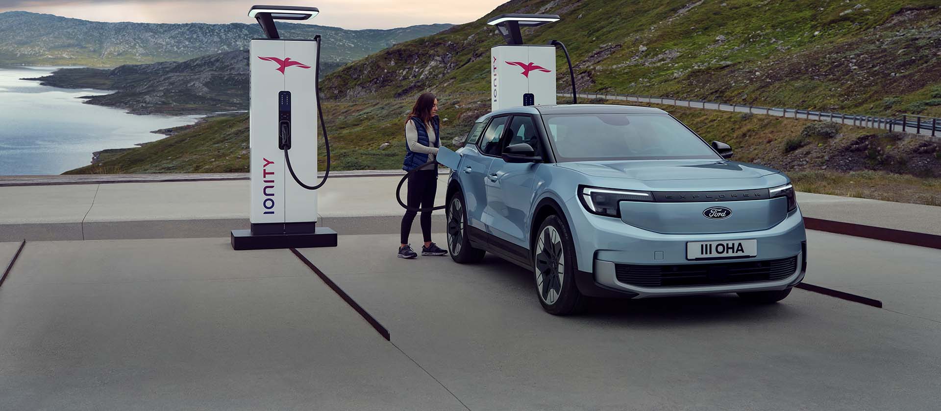 O femeie conectează un Ford Explorer albastru complet electric, la o stație publică de încărcare folosind un cablu. Mașina este parcată lângă munți și apă.