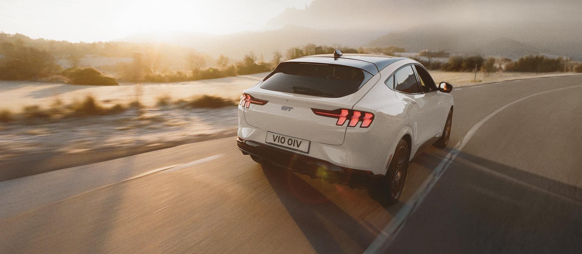 Un vehicul electric Ford Mustang Mach-E argintiu care conduce pe un drum lung deschis.