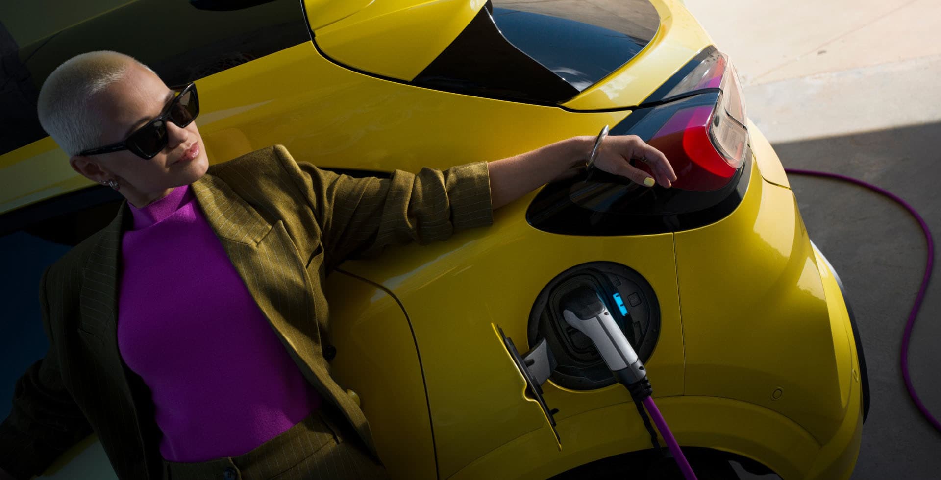 O femeie sprijinită de un Ford Capri electric galben care este conectat la un încărcător pentru vehicule electrice.