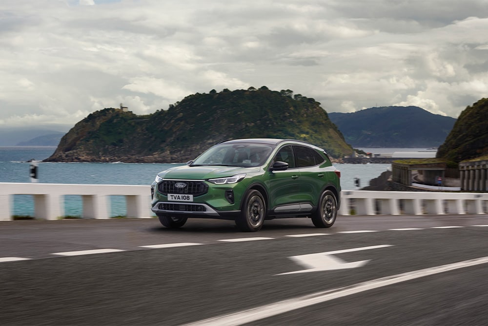 O mașină hibridă Ford Kuga verde cu încărcare automată care conduce de-a lungul unui drum pitoresc.