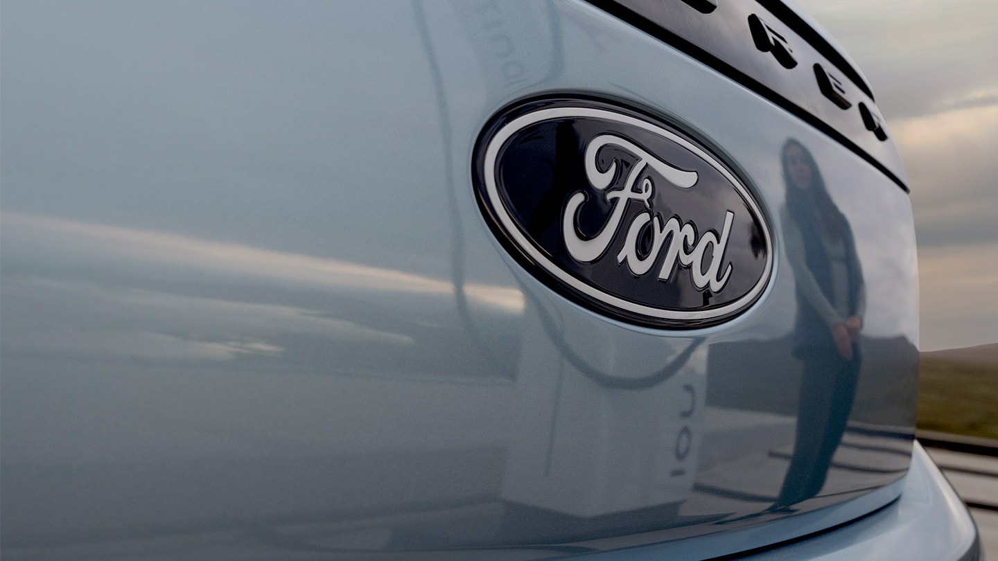 Sigla Ford pe un Ford Explorer electric albastru cu o stație publică de încărcare reflectată în mașină.