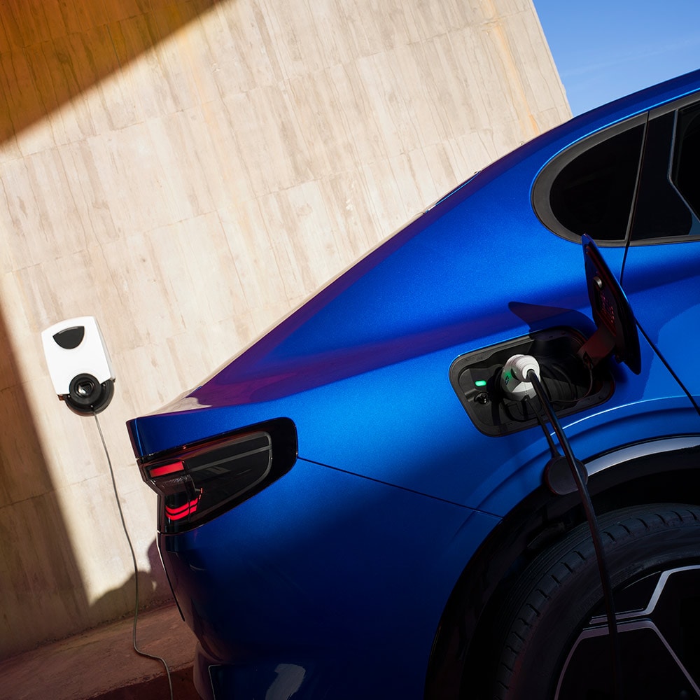 O vedere din spate a unui vehicul electric Ford Capri de culoare albastră conectat la un punct de încărcare de acasă pentru vehicule electrice.