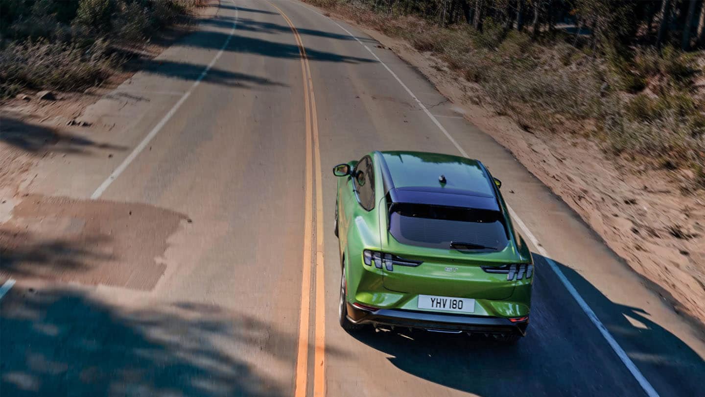 Un vehicul Ford Mustang Mach-E GT în culoarea Eruption Green deplasându-se pe un drum de munte mărginit de copaci.