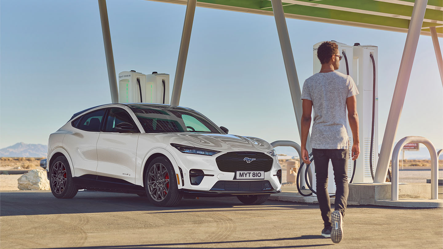 Un vehicul Ford Mustang Mach-E alb parcat într-o stație de încărcare publică în aer liber. Un bărbat în tricou alb se îndreaptă spre mașină.
