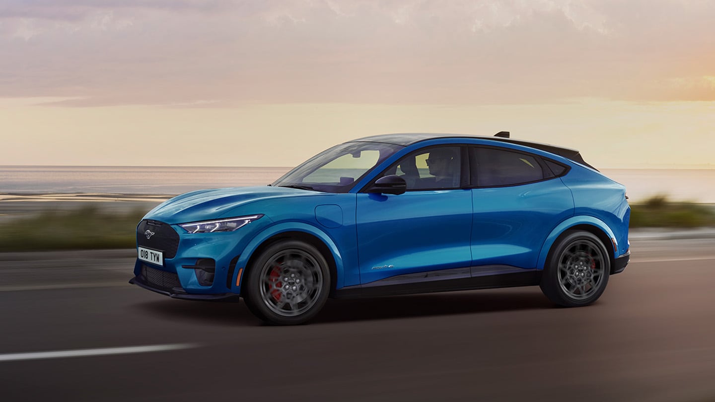 O imagine laterală cu un model Ford Mustang Mach-E electric albastru care se deplasează pe un drum de coastă, la apus.