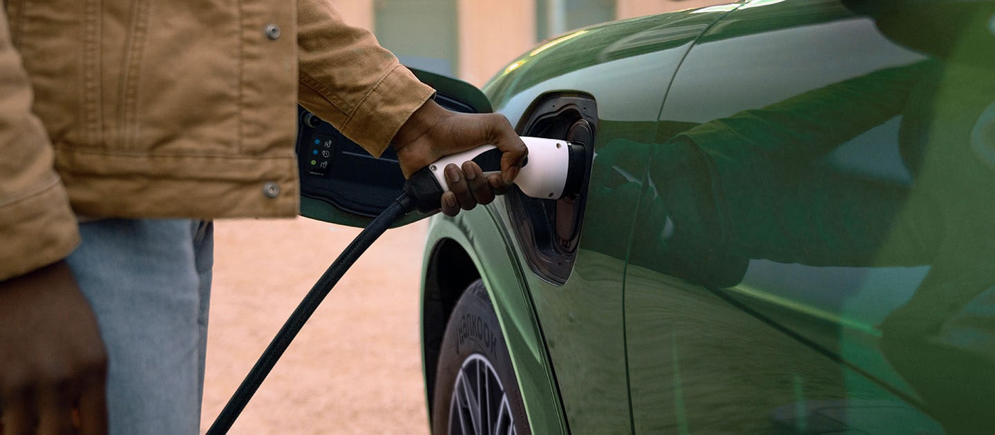O persoană într-o jachetă crem care conectează un cablu de încărcare electrică la vehiculul său Ford Mustang Mach-E electric, verde.