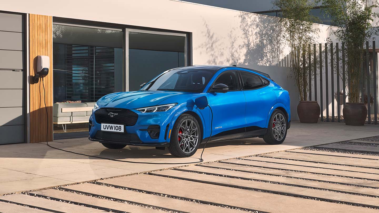 Un vehicul Ford Mustang Mach-E electric albastru conectat prin cablu la un încărcător auto pentru acasă în timp ce este parcat pe alee langă o casă modernă.
