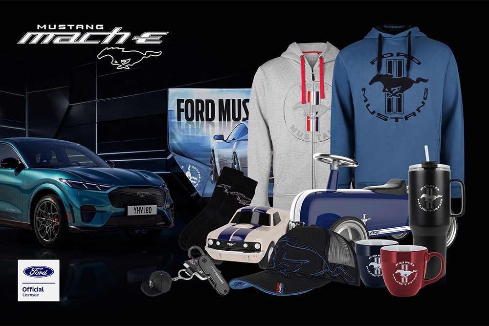 Colecție de produse promoționale cu diverse vehicule Ford în fundal, cu umbrelă, căni și articole de îmbrăcăminte cu marca Ford.