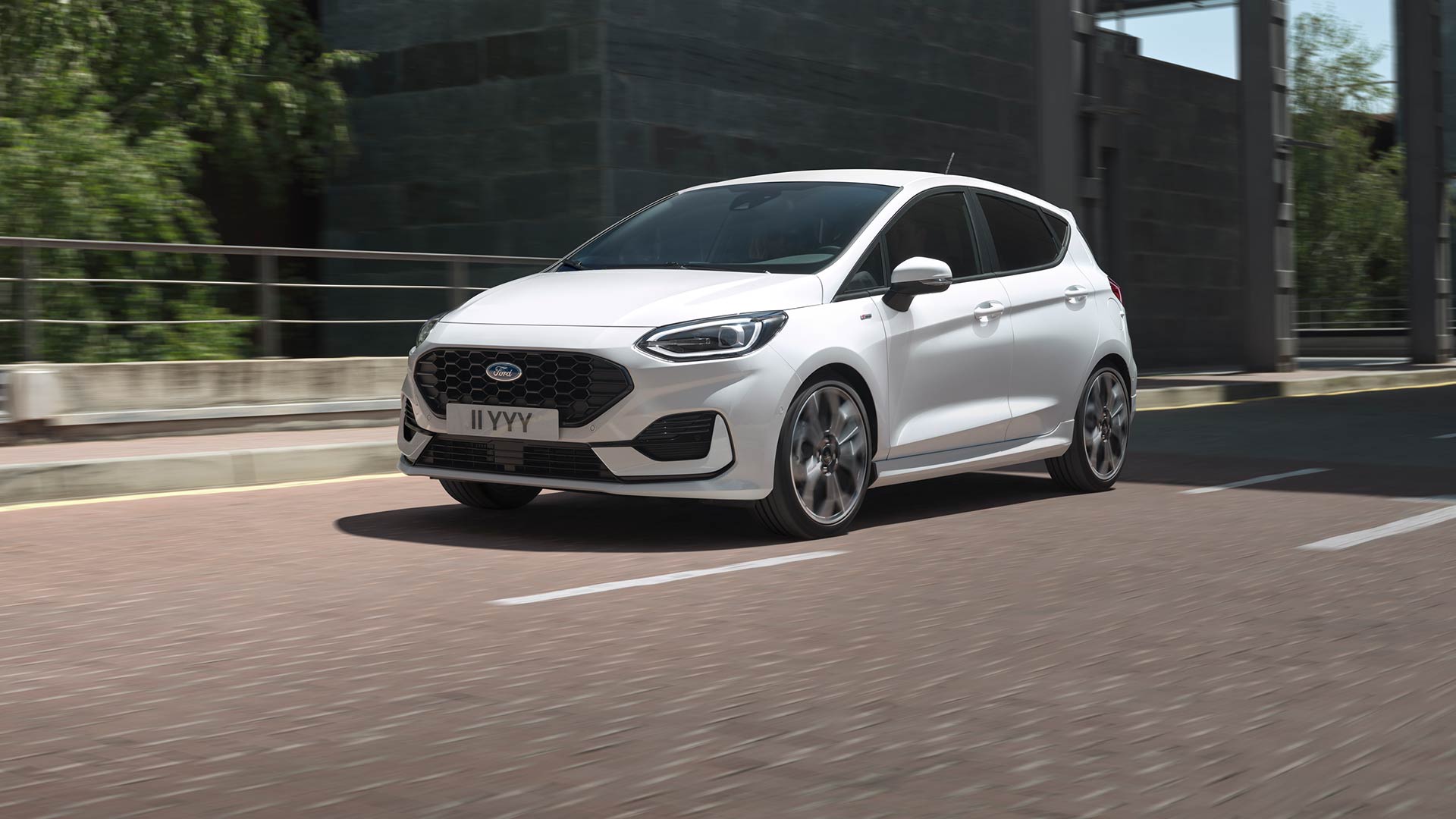 Un Ford Fiesta alb care rulează pe un drum urban, cu vegetație și clădiri în fundal.
