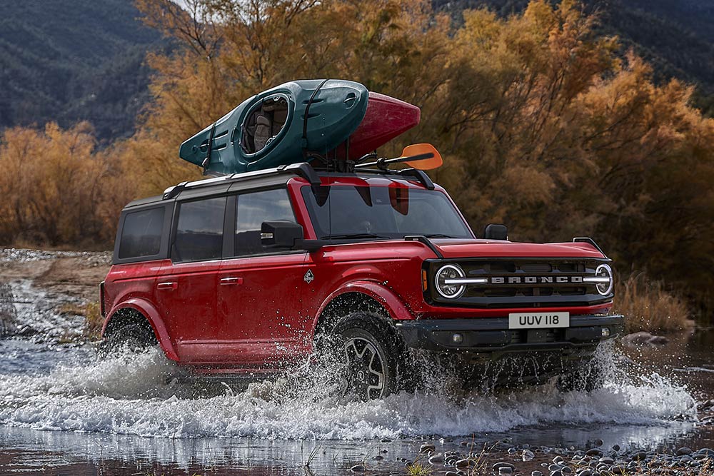Vedere din lateral a unui model Ford Bronco roșu deplasându-se prin apă puțin adâncă într-un cadru montan.