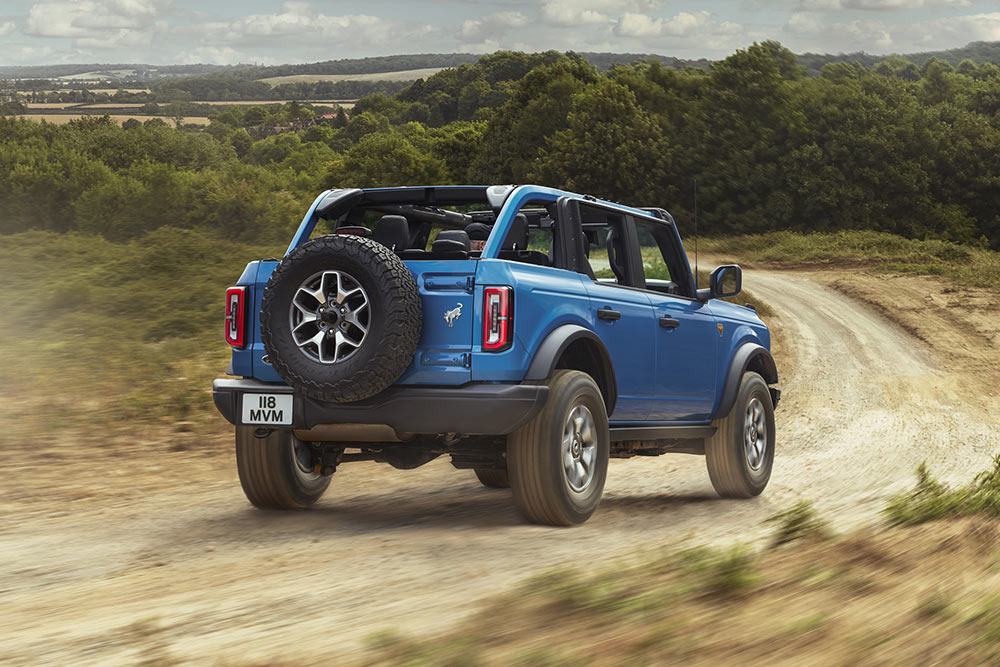 Vedere din spate a unui model Ford Bronco albastru deplasându-se printr-un peisaj rural.