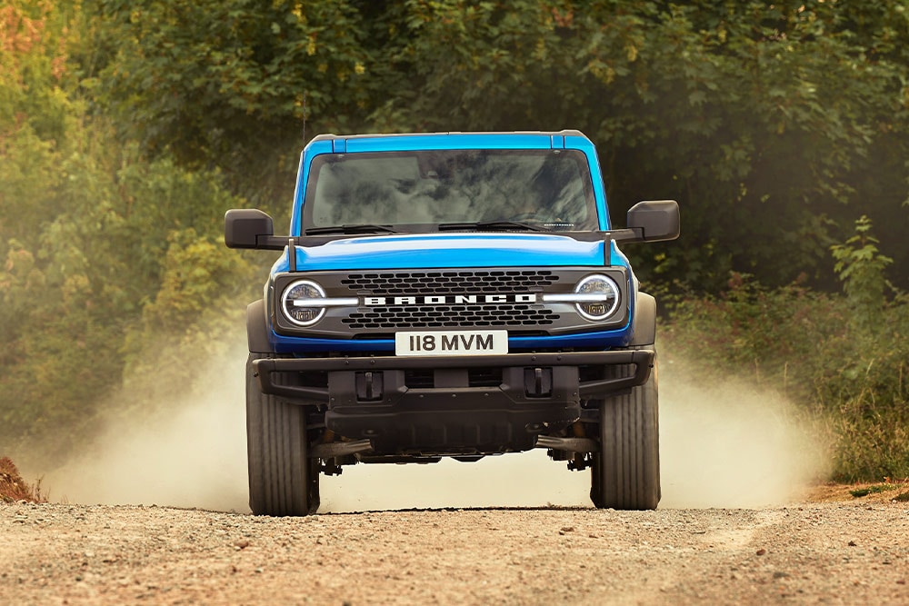 Vedere din față a unui model Ford Bronco albastru deplasându-se pe un drum de pământ. 