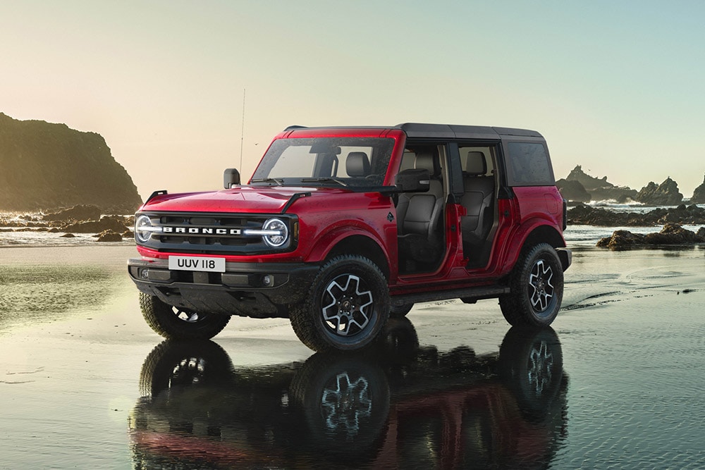 Vedere din lateral a unui model Ford Bronco roșu, cu ușile demontate, parcat pe un mal nisipos umed.