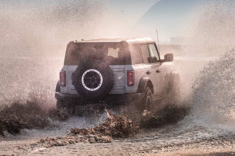 Vedere din spate a unui model Ford Bronco argintiu deplasându-se printr-un jet de apă pe o plajă.