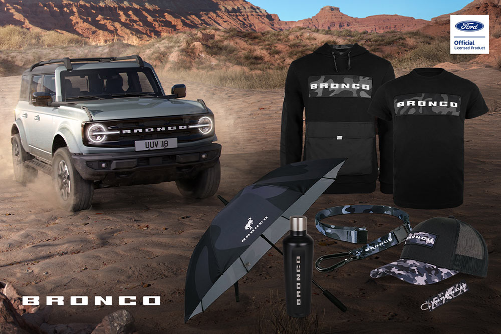 Colecția Ford Bronco, incluzând un tricou negru, o jachetă, un tricou polo, o șapcă, o căciulă și o geantă de voiaj, cu un model Bronco gri în fundal.
