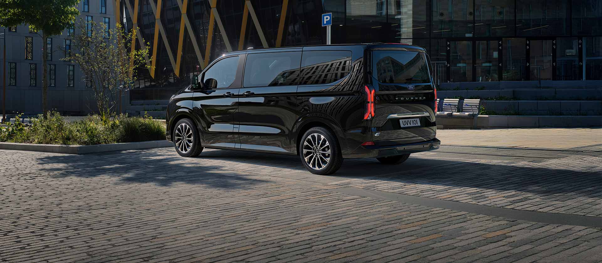 Un Ford E-Tourneo Custom negru este parcat în fața unei clădiri moderne, prezentând partea laterală și partea din spate.