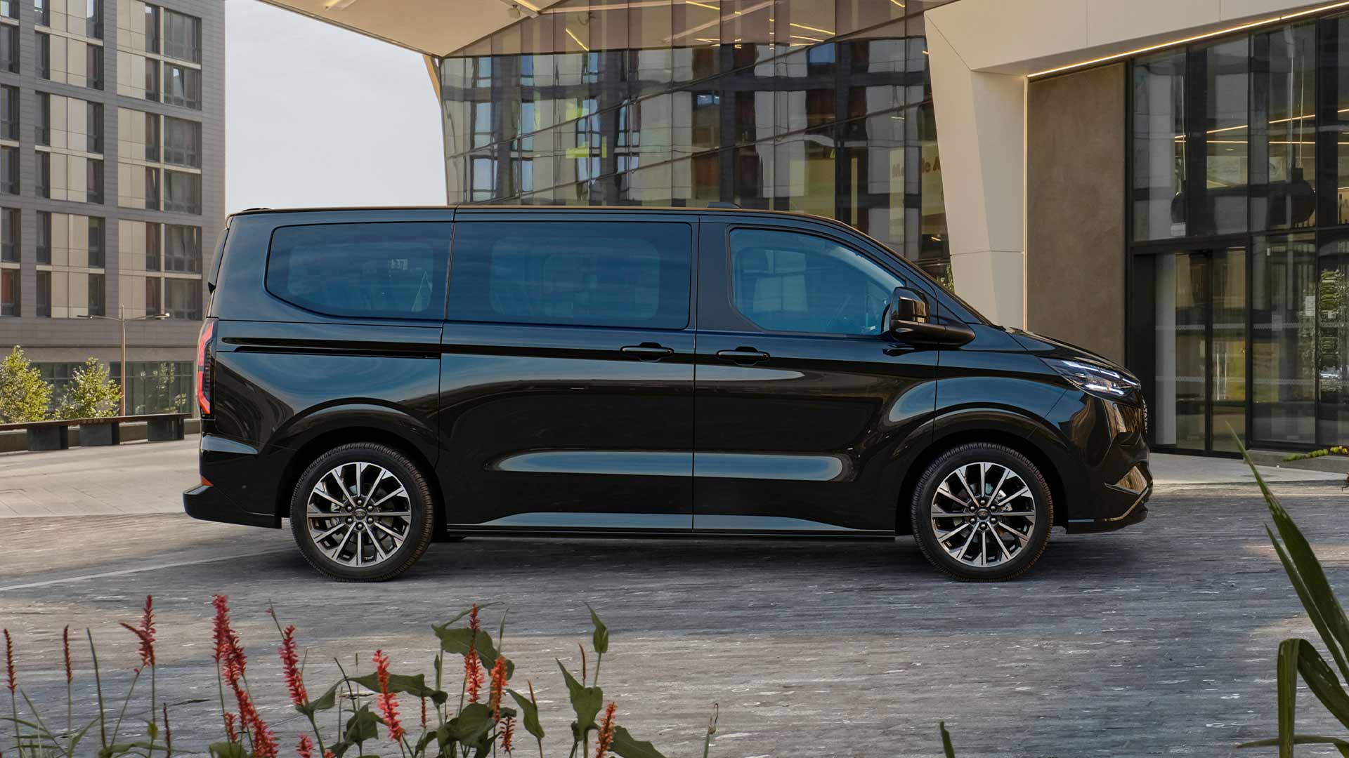 Un Ford E-Tourneo Custom negru este parcat în fața unei clădiri moderne.