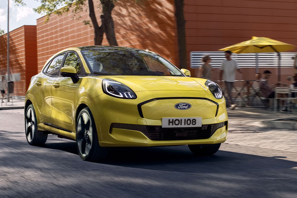 Vedere frontală a unui model Ford Puma Gen-E® electric, de culoare galben, care circulă pe o stradă dintr-un oraș.