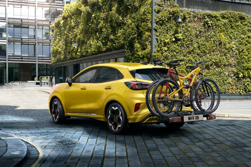 Un Ford Puma Gen-E galben parcat într-un cadru urban, cu două biciclete montate pe un suport de biciclete în spate.