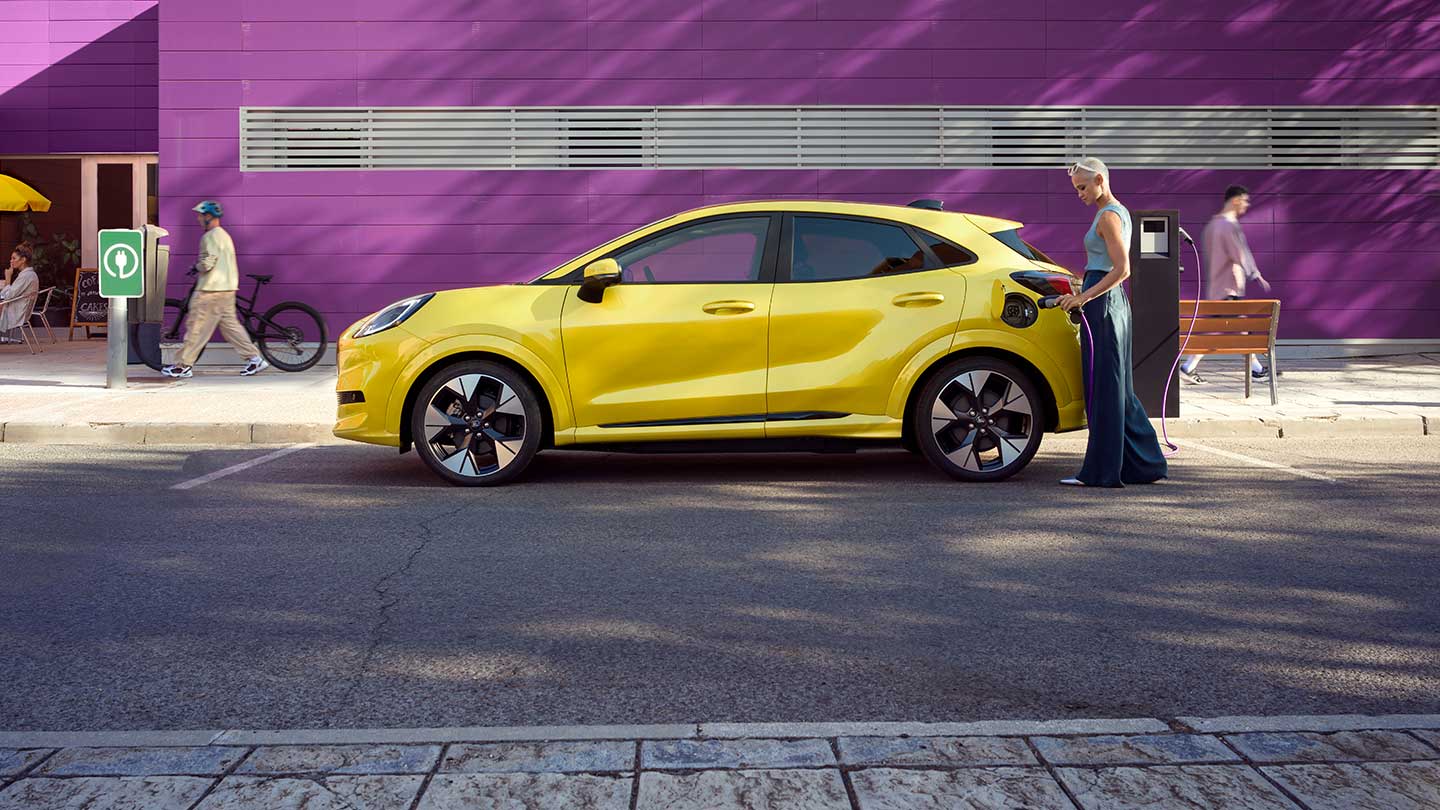 Vedere laterală a Ford Puma Gen-E® galben, conectat la o stație de încărcare publică, în timp ce se află în parcare.