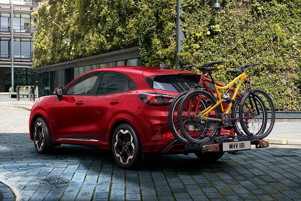 Un Ford Puma Gen-E galben parcat într-un cadru urban, cu două biciclete montate pe un suport de biciclete în spate.
