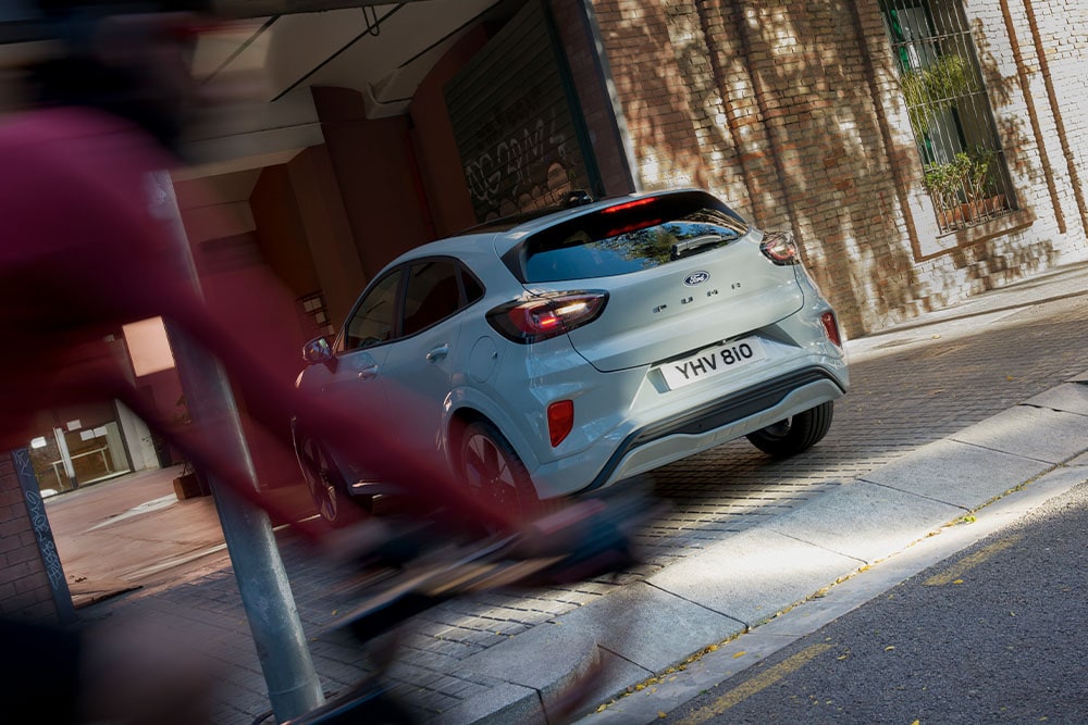 Vedere din spate a unui vehicul Ford Puma  gri parcat pe o stradă urbană cu vegetație.