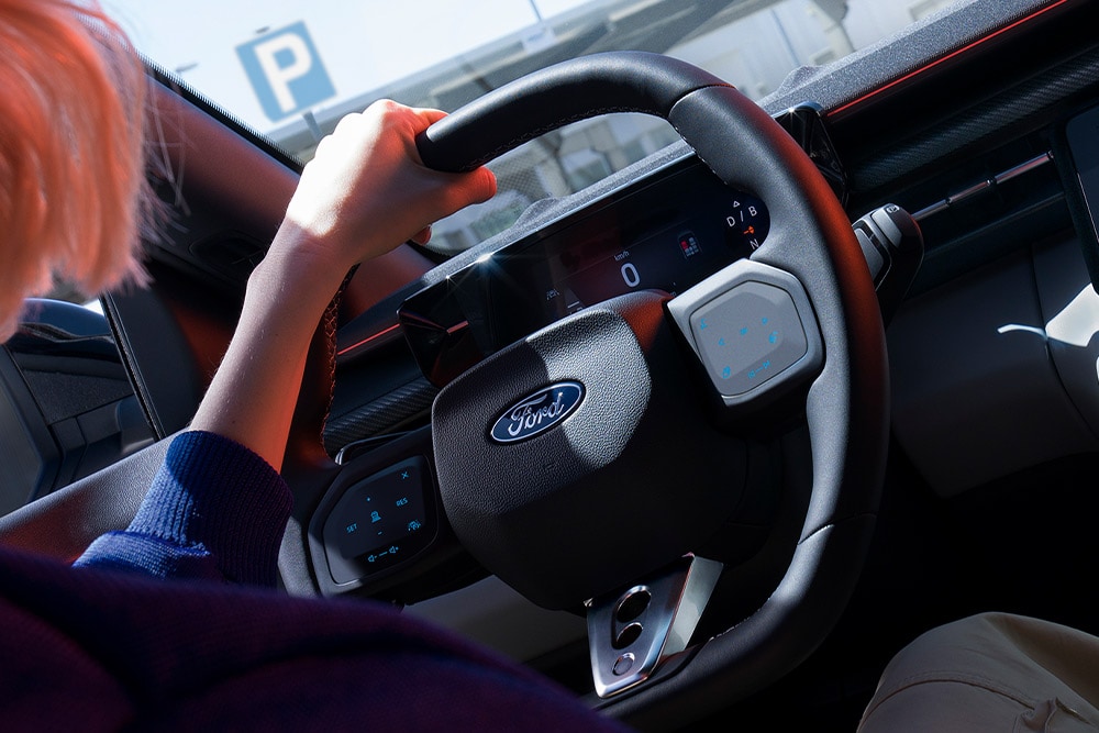 Un prim-plan cu o femeie care ține mâna pe volanul unui Ford Capri parcat, imaginea arătând și clusterul digital de instrumente.