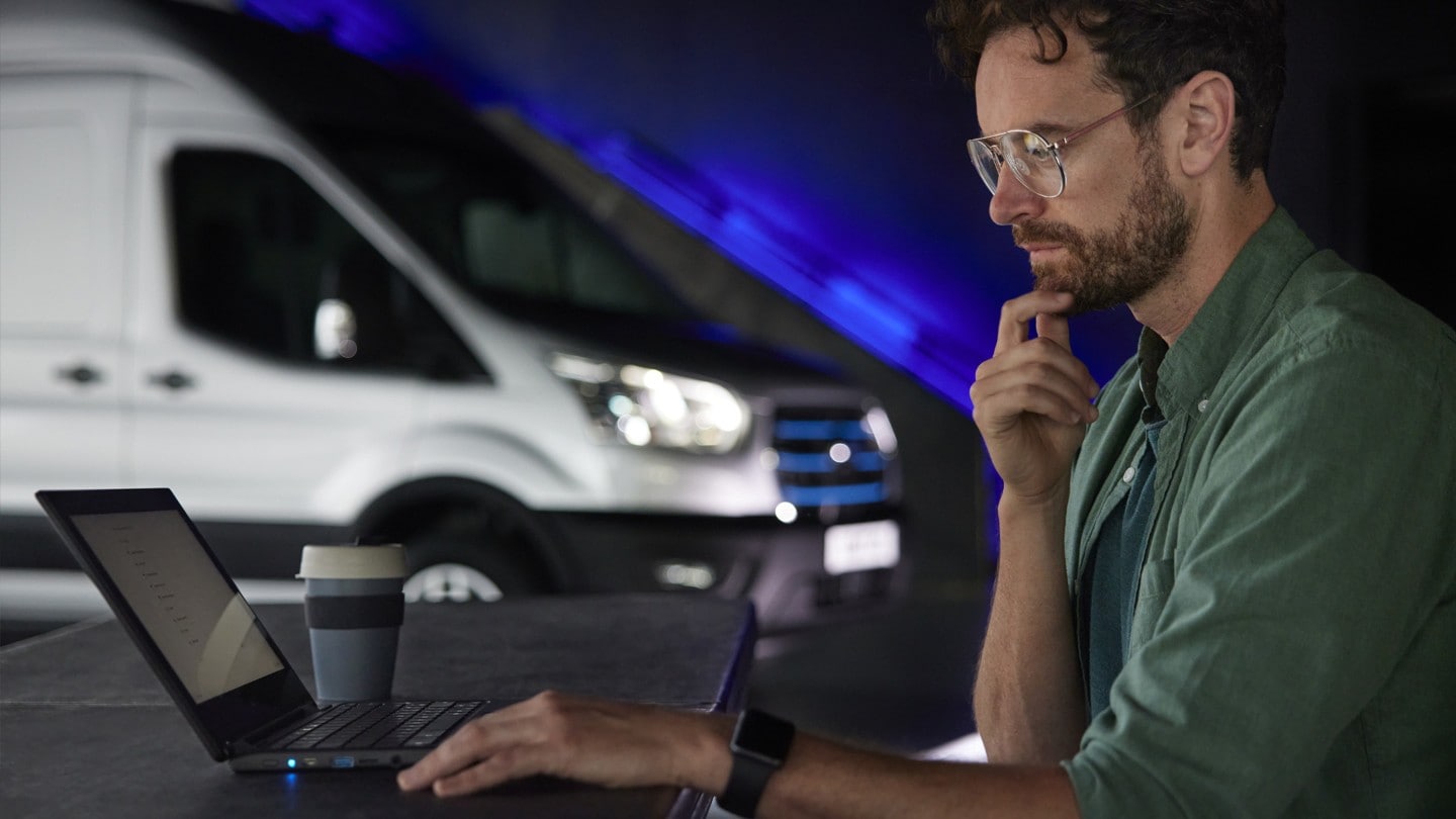 Persoană care lucrează pe laptop cu Ford E-Transit în fundal