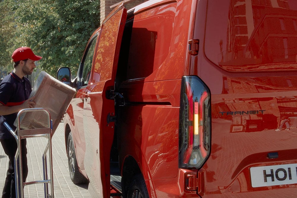Curier care descarcă colete dintr-un Ford Transit Custom culoare roșie.