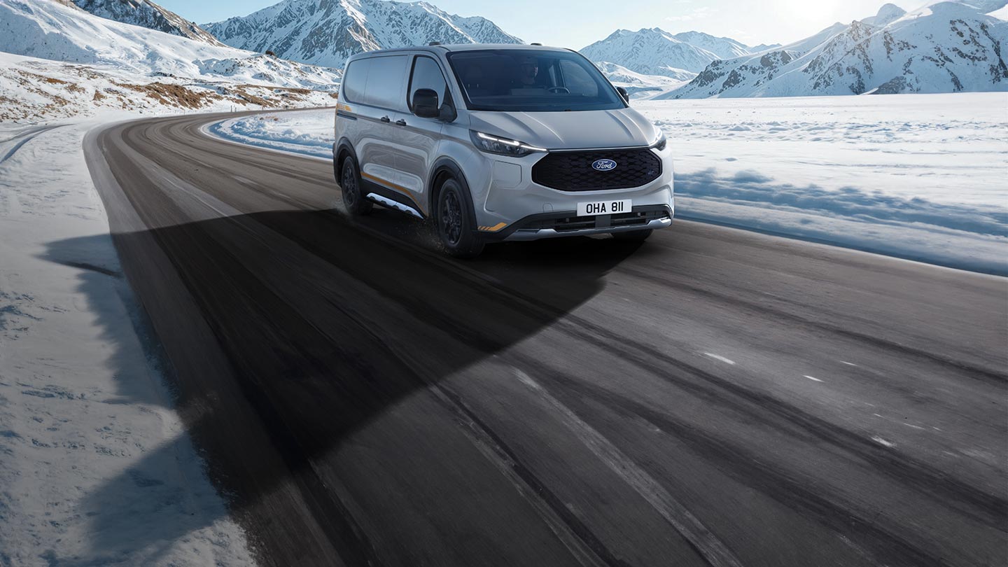 A silver Ford E-Transit Custom driving on a snowy mountain road under a clear blue sky.