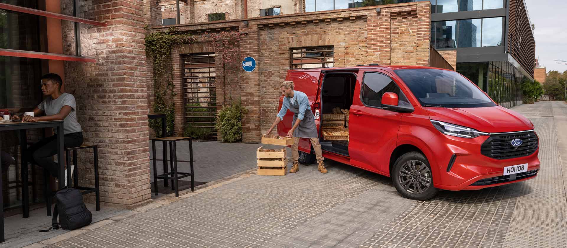 Furgon Ford Transit Custom  culoare roșie parcat în fața unei cafenele.