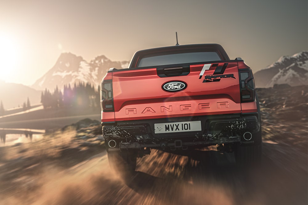 Vedere din spate a unui Ford Ranger Raptor roșu, care se deplasează pe un drum de pământ, cu munți în fundal.