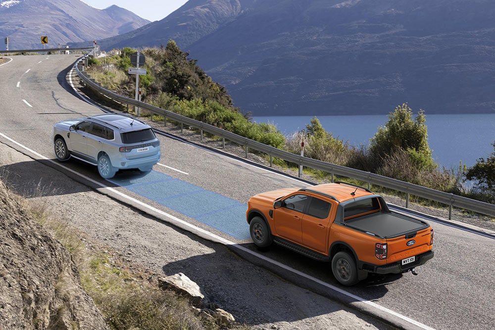 Ford Ranger Raptor culoare Code Orange, care se deplasează de-a lungul unui traseu montan pitoresc, evidențiind sistemele de asistență a șoferului.