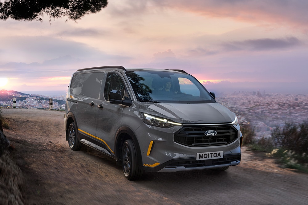 Grey Ford Transit Custom Trail van with yellow accents drives on rough road.