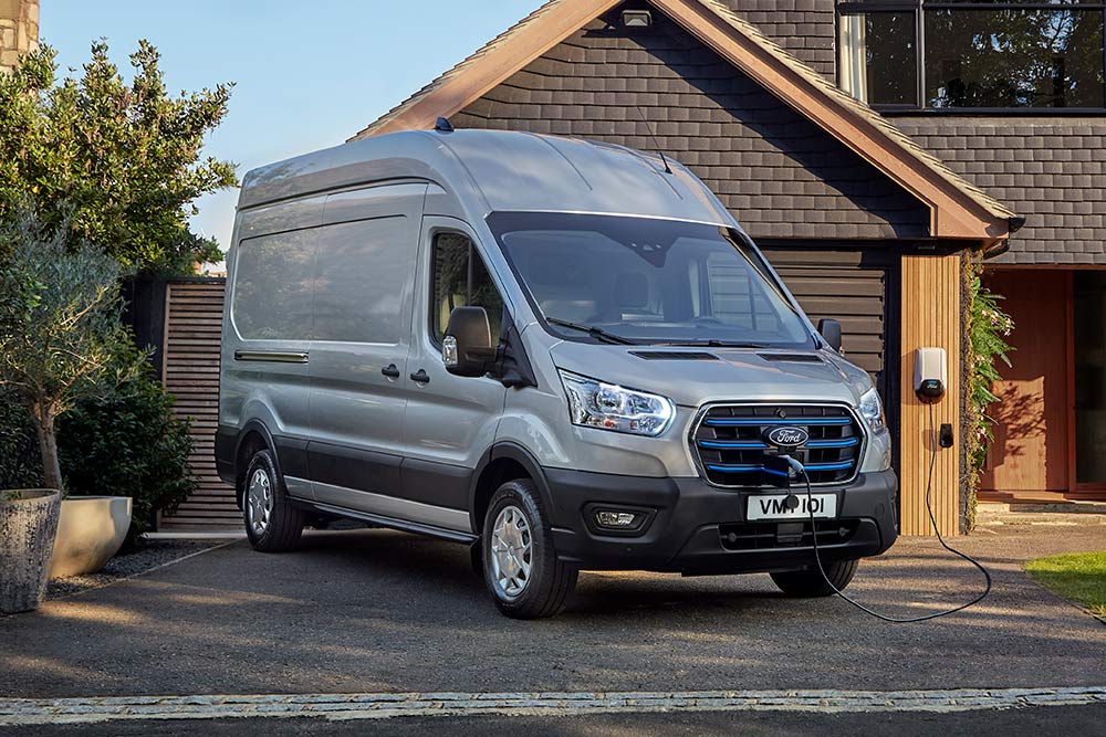  Ford E-Transit Furgon, Moondust Silver parcat pe o alee de lângă o casă, conectată la o stație de încărcare.
