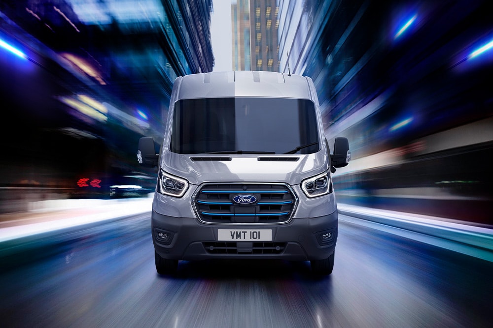 Vedere din față a două Ford Transit Furgon  parcate într-un garaj cu priveliștea unui oraș văzută prin ferestre.