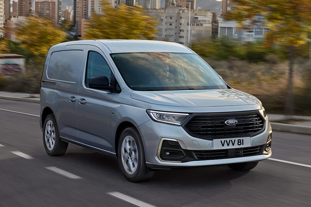 Un Ford Transit Connect deplasându-se într-un oraș, prezentând ramele cromate ale proiectoarelor de ceață și farurile automate.