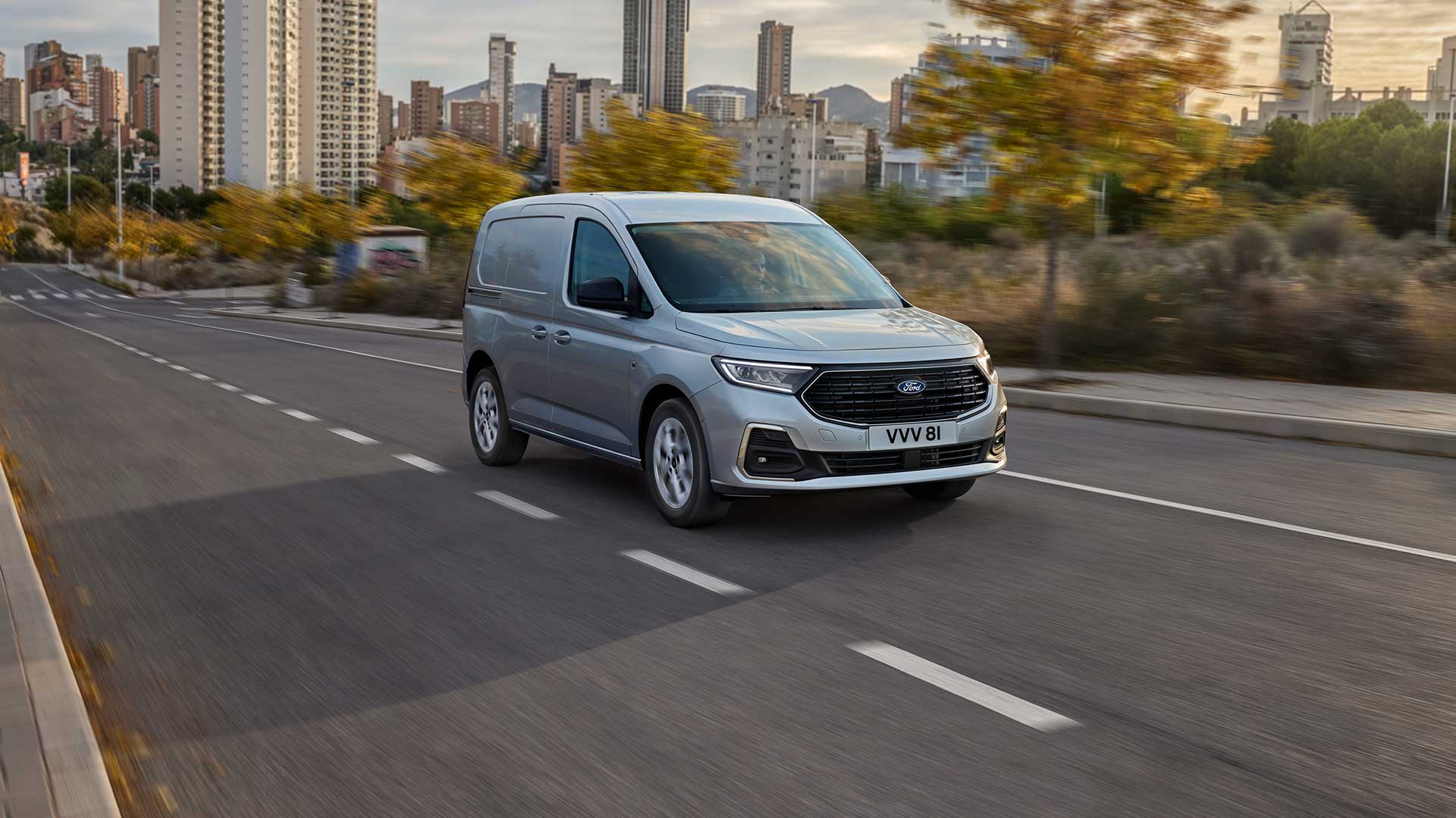 Un Ford Transit Connect deplasându-se într-un oraș, prezentând ramele cromate ale proiectoarelor de ceață și farurile automate.