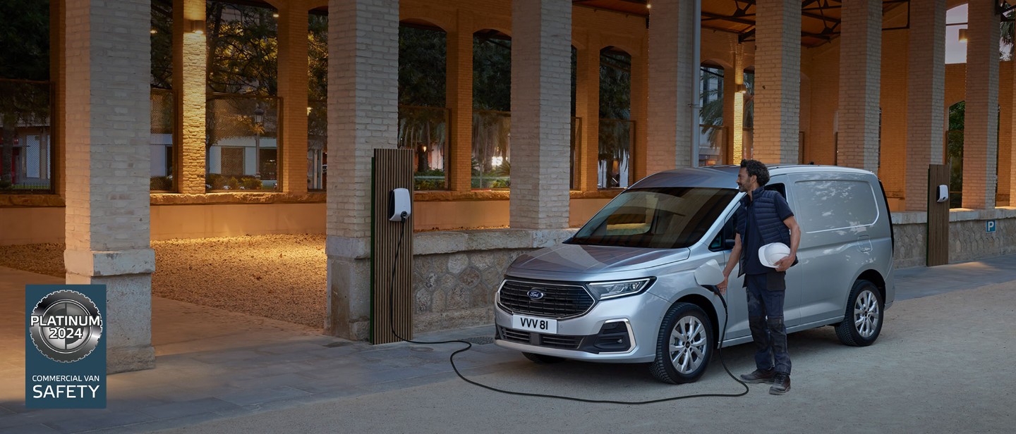 Un bărbat care ține o cască din material tare conectează un cablu de încărcare a cutiei de perete la noul Ford Transit Connect în culoarea stardust silver.