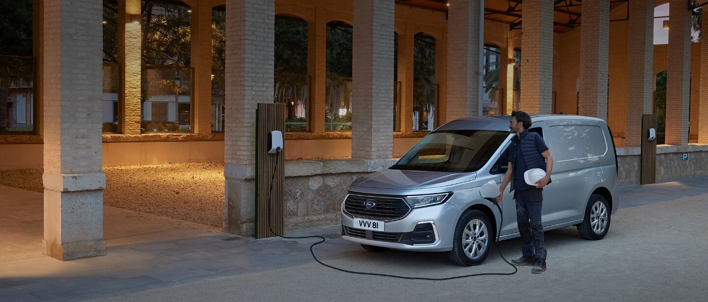 Un bărbat care ține o cască, conectează un cablu de încărcare la priză un Ford Transit Connect în culoarea Stardust Silver.
