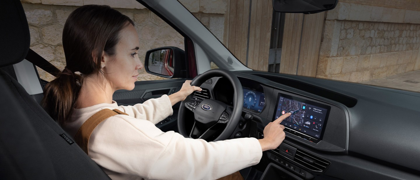 O femeie atinge ecranul tactil de 10” [25 cm] în timp ce stă pe scaunul șoferului al unui model nou Ford Transit Connect.