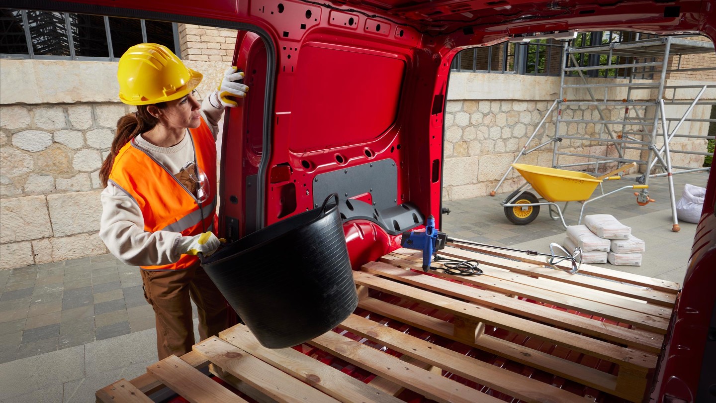 O femeie pune materiale în spațiul de încărcare al noului Ford Transit Connect în culoarea Maple Red, cu ușile laterale și din spate deschise, pe un șantier de construcții.