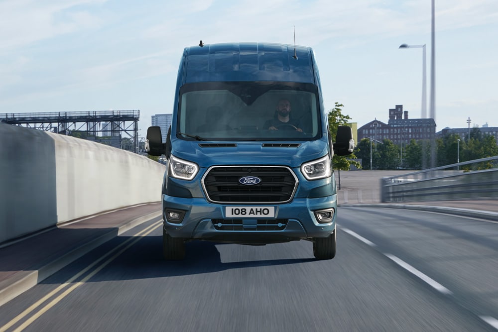 Vedere din față a unei furgonete Ford Transit albastre care circulă pe un drum. 