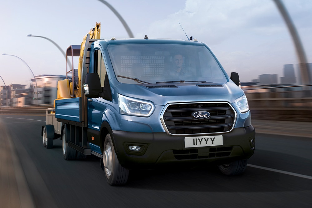 Vedere frontală a grilei unui Ford Transit autoșasiu cu cabină care transportă un excavator în timp ce se deplasează pe un drum urban. 