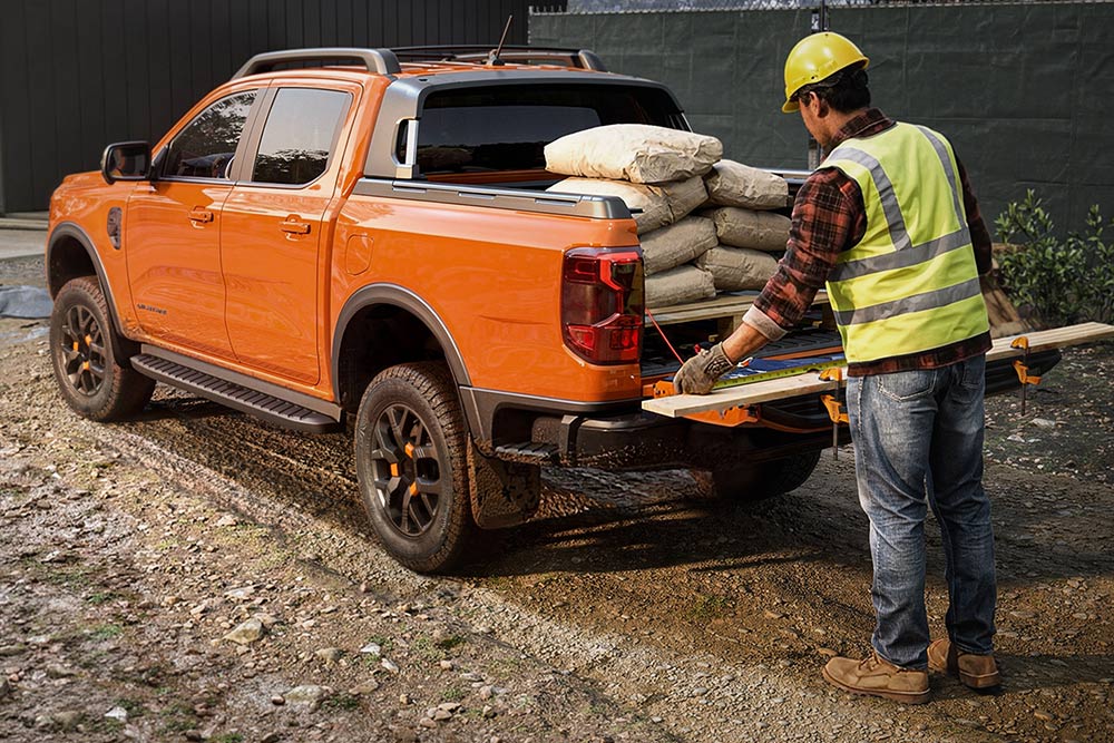 Un muncitor în construcții care încarcă echipamente pe un pat portocaliu al unui vehicul Ford Ranger.