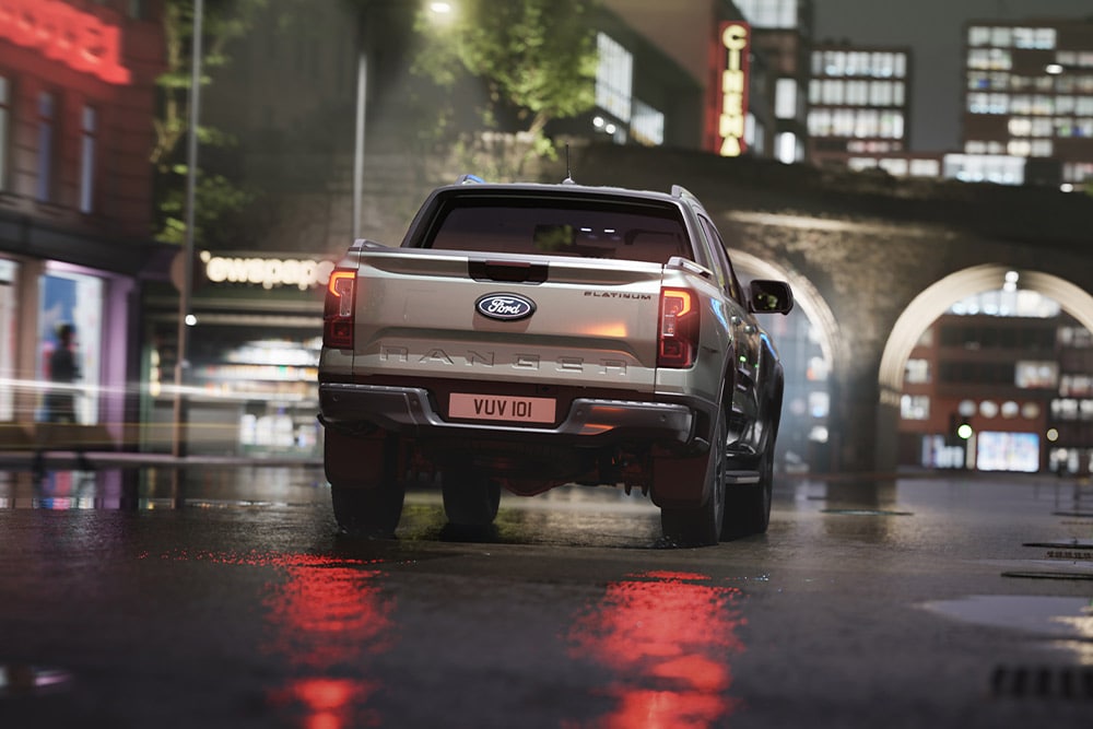 Vedere din spate a unui Ford Ranger argintiu care este condus printr-un peisaj urban luminat de neon pe timp de noapte.