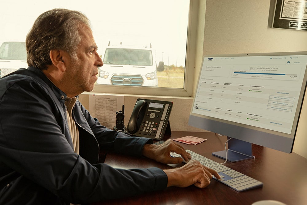 O persoană la birou privește un monitor, cu o dubă Ford albă vizibilă prin fereastră.