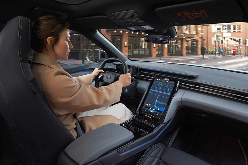 O persoană care conduce un vehicul Ford, cu ecranul tactil central mare care afișează navigația.