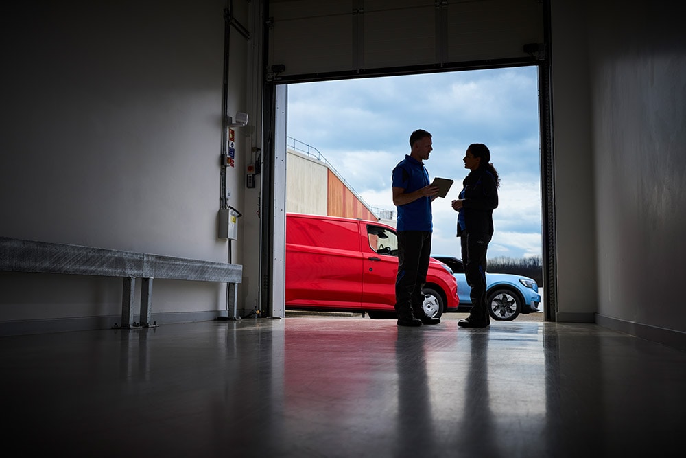 Două persoane care discută în dreptul unei uși de garaj, cu o furgonetă Ford roșie și un SUV Ford albastru parcate afară.